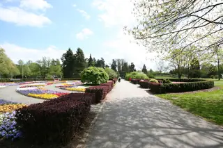 A paved path runs between clipped hedges and colorful flower beds, with trees and a few distant walkers.