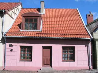 Small pink house with a steep red-tile roof, dormer window, brown door and two street-level windows.