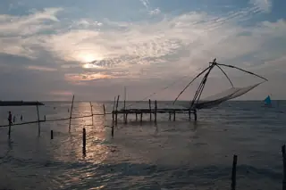 A large cantilevered fishing net hangs above the water beneath a clouded sunset, with wooden platforms and a small figure at the left edge.