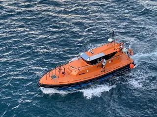 An orange working boat crossing blue water with a person standing beside its cabin.