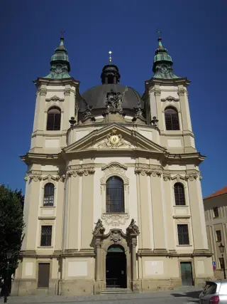 Cream-colored Baroque church facade with two green-capped towers and sculpted figures above its doorway.