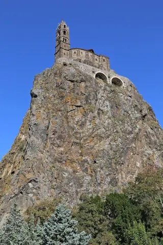 A stone chapel and bell tower stand on a steep rocky pinnacle against a clear blue sky.
