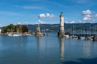 A stone lion and lighthouse stand across the harbour entrance, with moored sailboats, rippling blue water and distant wooded hills.