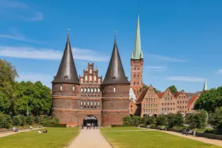 Twin round brick gate towers face a lawn and path, with gabled warehouses and a tall church spire behind.