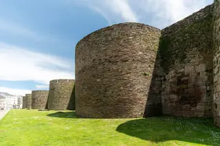 Rounded stone towers and a long defensive wall rise beside a grassy strip under a blue and white sky.