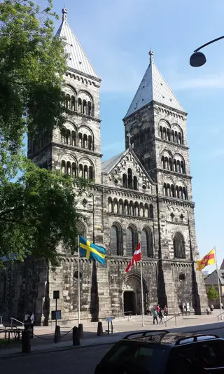 Two stone church towers rise above an ornate entrance, with leafy trees, flags and a few small visitors below.