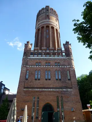 Tall round brick water tower with small corner turrets and arched windows, beneath blue sky.