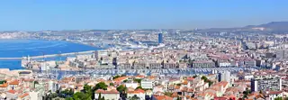 A wide city panorama extends around a harbour filled with boats, with the sea and distant hills beyond.