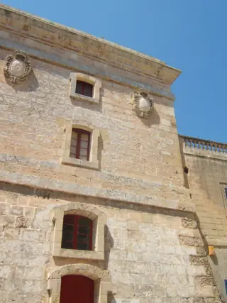 An upward partial view of a limestone tower facade with red-framed windows and two carved heraldic shields.