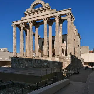 Tall stone columns and a partial pediment before an older palace facade, with low modern walkways around the temple.