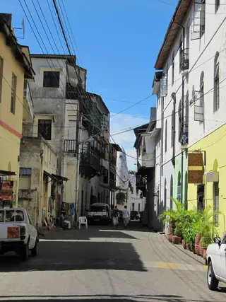 Narrow street with projecting balconies, overhead wires, parked vehicles and two people walking away.