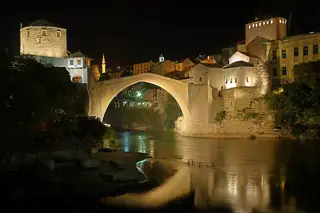 A floodlit stone arch spans a dark river, with warm reflections beneath stone towers and riverside buildings.
