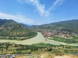 Two broad rivers meet between green hills and tree-lined banks, with Mtskheta’s rooftops in the distance.