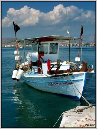 White and blue fishing boat with anchors and ropes moored beside a quay, with distant waterfront buildings.