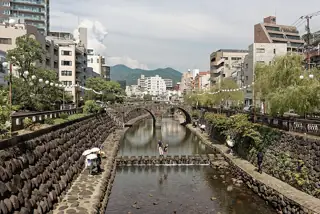 A twin-arched stone bridge crosses a narrow river between planted urban banks, with people and parasols along the lower paths.