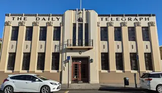 Cream Art Deco facade with tall dark windows and parked cars at street level.