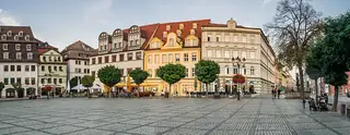 Wide view across a cobbled square toward colorful merchant houses, café tables and trimmed trees.