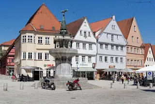 An eagle-topped stone fountain stands before gabled buildings, with parked motorcycles and café visitors around the square.