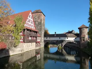 Half-timbered riverfront building and stone tower beside a covered arched bridge reflected in blue water.
