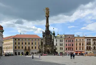 Holy Trinity Column amid the paving and buildings of Horní náměstí, with visitors beneath a cloud-filled sky.