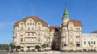Broad cream-colored palace facade with green-roofed towers, cafe parasols and a sunny square.
