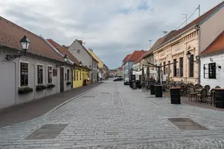 A broad paved street runs between pale historic facades, with café chairs along the right side.