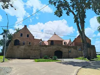 Brick buildings and a stone defensive wall around an arched fort entrance, with a large tree beside the road.