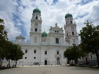 Two green-domed towers and a broad white church facade rise behind trees and a paved square.