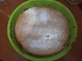 Round golden cake dusted with powdered sugar in a green baking mould on a wooden table.
