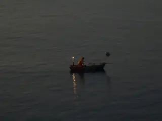 A small dark fishing boat with one fisherman and a glowing lamp reflected in the surrounding dim sea.