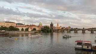 Stone arches and riverside buildings reflected in the Vltava, with boats beneath a broad cloudy sky.