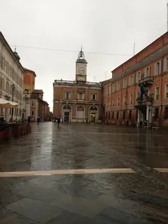 Umbrella-carrying walkers cross wet paving toward the clock-fronted buildings of Piazza del Popolo.