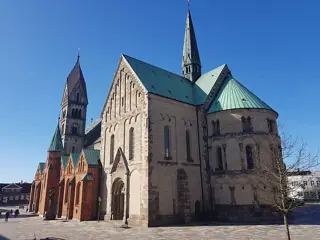 Stone cathedral apse and gabled transept with copper roofs, a small bronze monument and blue sky.
