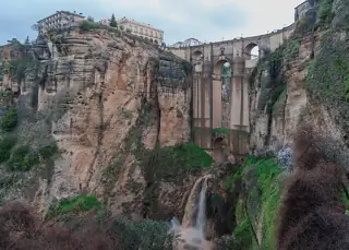 Stone bridge spanning a deep rocky gorge, with cliff-top buildings and a waterfall below.