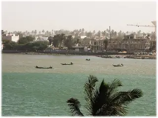 Small boats cross green river water between a foreground palm and a hazy construction-lined bank.