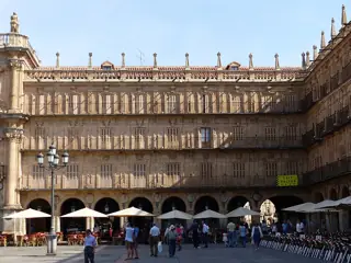 Stone arcades and rows of balconies above cafe parasols and a group of pedestrians in a sunlit square.