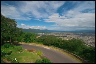 A winding hill road and green trees overlook a distant city beneath mountains and a broad clouded sky.