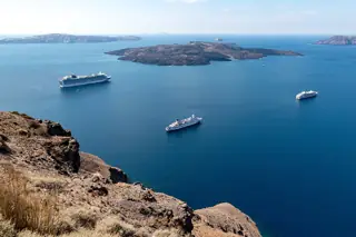 Blue caldera water and a dark volcanic island beyond a dry rocky foreground, with three cruise ships and distant ridges.