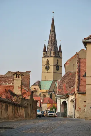 Patterned pointed church tower above tiled roofs and a cobbled lane, with scaffolding and green roof covering below.