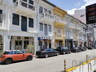 White and yellow shophouse facades with shuttered windows and balconies above a row of parked cars.