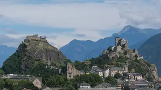 Two rocky hills crowned by historic buildings rise above town rooftops, with layered mountains and clouds behind.