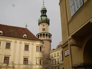 A green-copper tower with a clock and arcaded balcony rises between decorated yellow buildings beneath a grey sky.