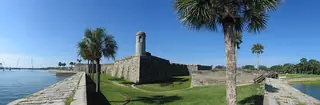 A wide panorama shows a stone bastion and watchtower beside the moat, palms and the bay seawall.