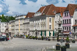 Broad paved square with ornamented historic facades, a sculpted fountain, cafe tables, parked cars and contextual pedestrians.