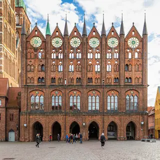 A brick town-hall screen facade with pointed pinnacles and circular decorations stands above people crossing a paved square.