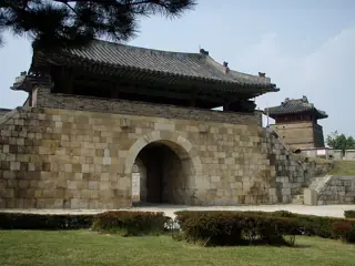 A tiled gate pavilion above a stone arch, framed by pine branches and hedges.
