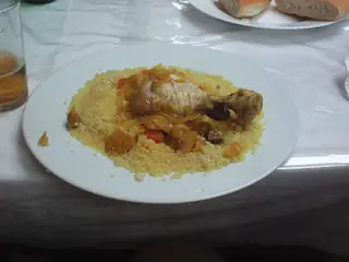 Chicken and vegetables over couscous on a plain white plate, with bread and a partly clipped drink.