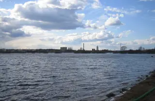 Broad river water beneath a cloudy sky, with a distant mixed city skyline and a clipped green rail at the edge.