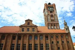 Gothic brick town hall and clock tower seen upward beneath bright clouds.