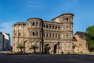 The Roman stone gate and its rounded towers rise beyond a broad road, with palms at the base and blue sky overhead.